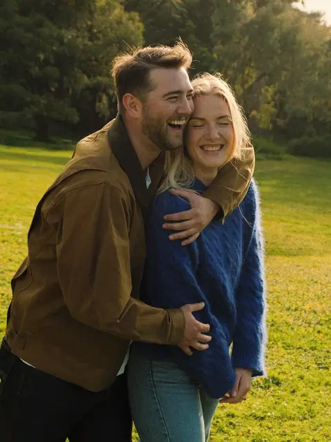Engagement photo on 35mm color film: a couple laughing with their heads pressed together in a golden-lit San Francisco park, he in a tan jacket with his arms wrapped around her blue cable-knit sweater.