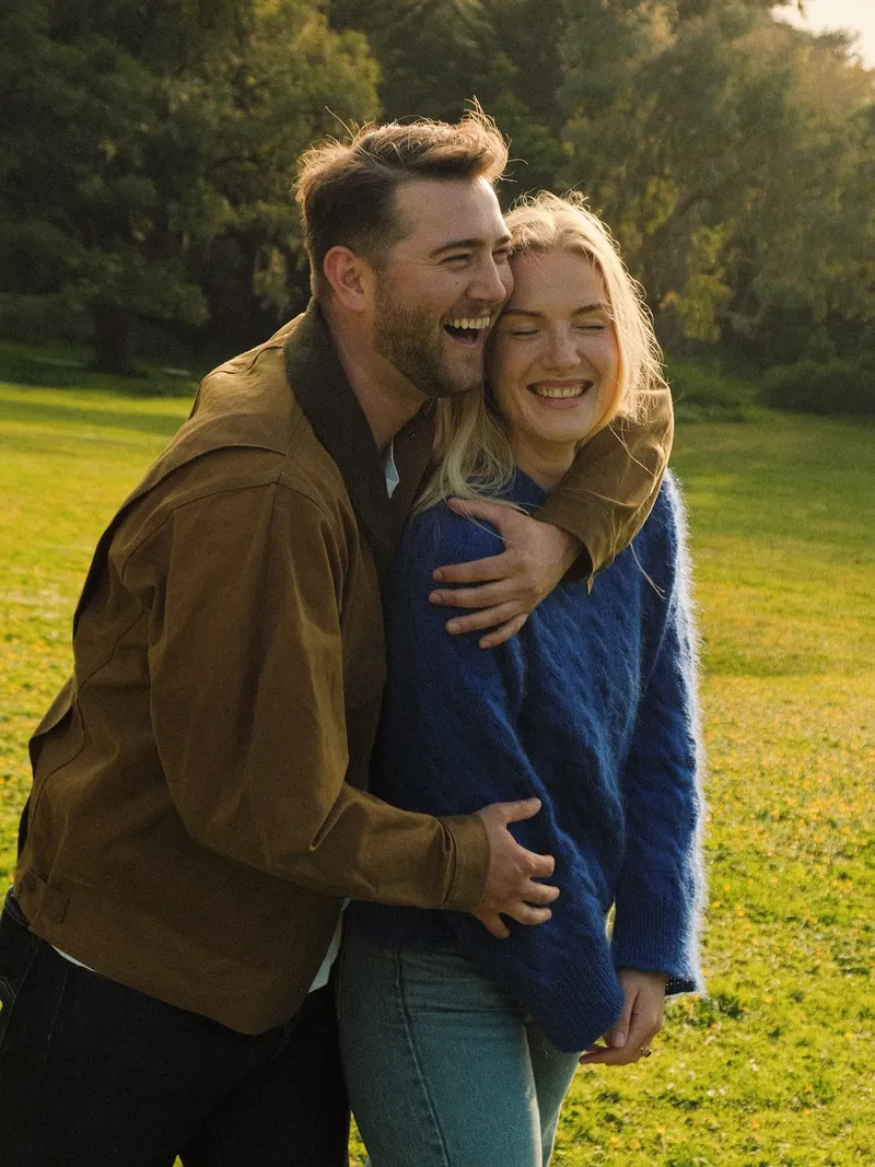 Engagement photo on 35mm color film: a couple laughing with their heads pressed together in a golden-lit San Francisco park, he in a tan jacket with his arms wrapped around her blue cable-knit sweater.