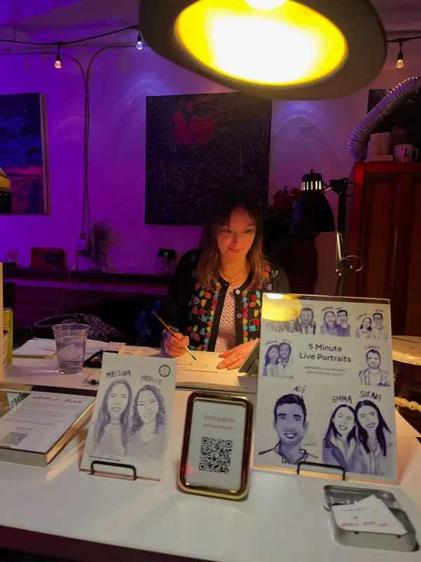 Natalie painting at her live portrait station, with a printed sign reading '5 Minute Live Portraits' and finished guest portraits propped on stands