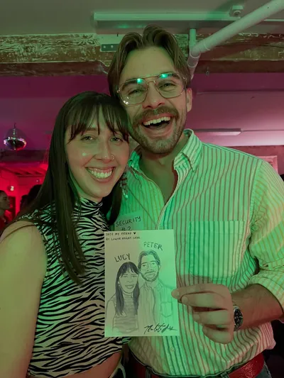 A couple beaming under pink and green party lights while holding the watercolor portrait Natalie painted of them