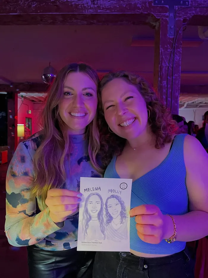 Two guests smiling under club lighting as they hold up the watercolor portrait Natalie painted of them at a Golden Gate Triathlon Club event
