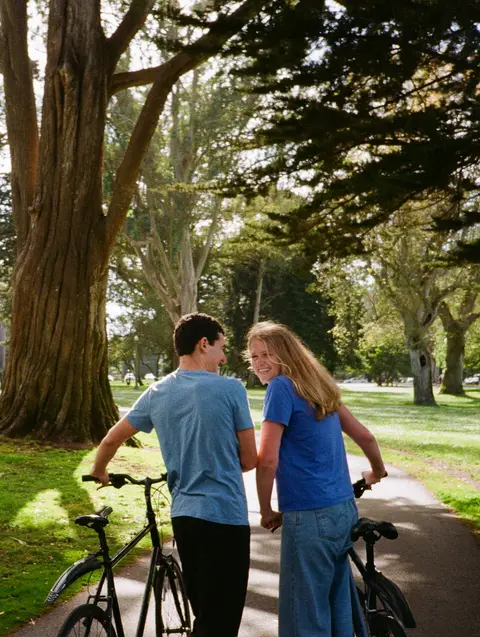 Engagement photo on 35mm color film of a couple walking their bicycles down a sunlit park path under a huge cypress tree, the woman glancing back over her shoulder and laughing.