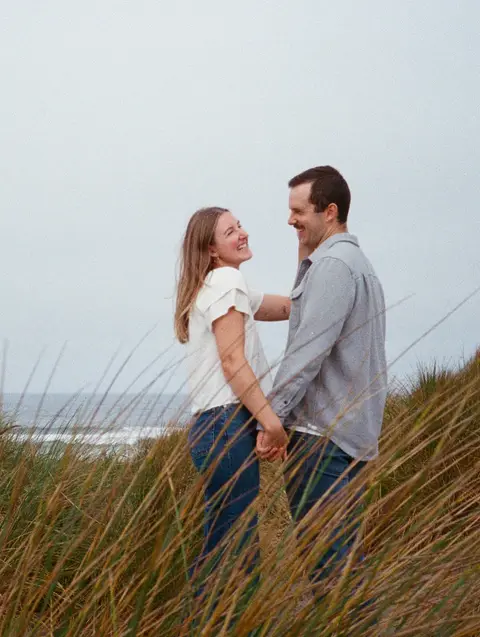 Engagement photo of a couple holding hands and laughing in tall dune grass above the ocean, shot on 35mm film.