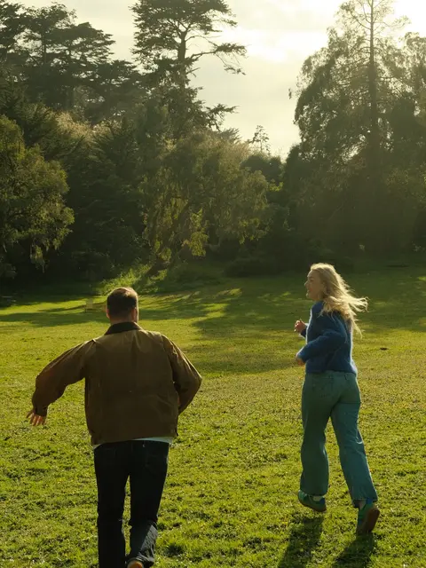 Engagement photo of a couple chasing each other across a sunlit meadow in a San Francisco park, shot on 35mm color film.