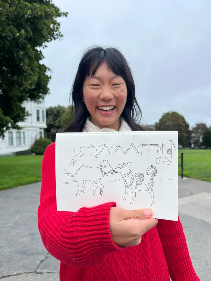A smiling participant in a red sweater holding up a sketchbook drawing at an outdoor workshop.