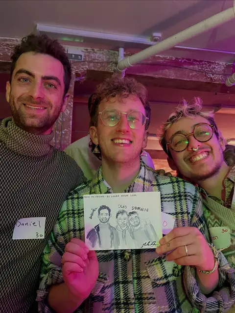 Three friends laughing together as they hold a live watercolor portrait of the three of them