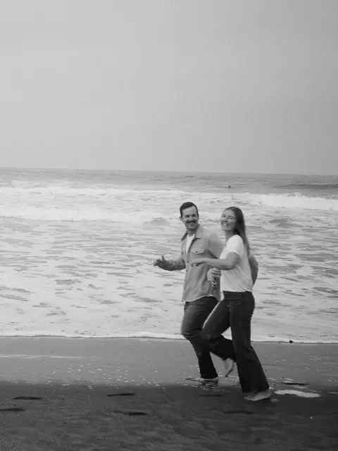 Black and white 35mm film photo of an engaged couple running along the waterline of a wide, empty beach, both grinning back at the camera.