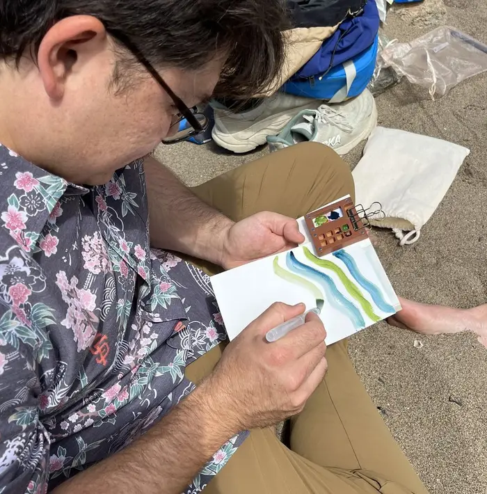 Overhead view of a participant sitting on the sand, mixing green and blue watercolor stripes in a small sketchbook with a travel palette clipped to the page.