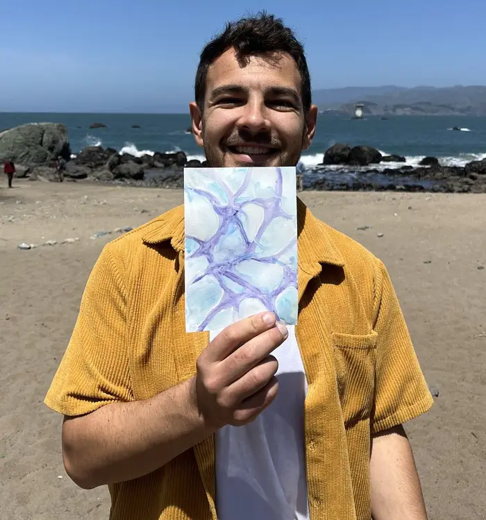 A participant in a mustard corduroy shirt smiling as he holds up a blue and violet watercolor study in front of the ocean at Mile Rock Beach.