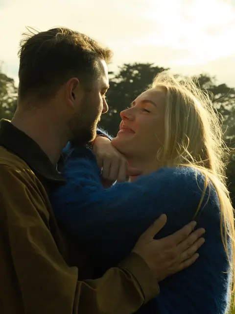Backlit engagement photo at sunset: a couple embracing with the low sun catching the edges of their hair, trees blurred behind them.