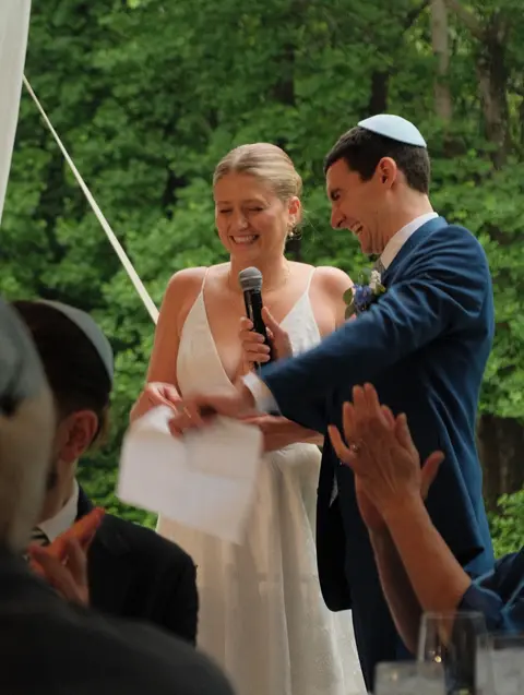 Wedding photo of a bride holding a microphone and laughing beside her groom during the ceremony at Wooden Sanctuary, guests applauding in the foreground.