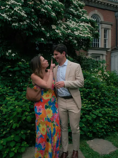 Two wedding guests joking with each other in front of a flowering dogwood and a brick building at the venue, she in a bright floral dress and he in a cream suit.