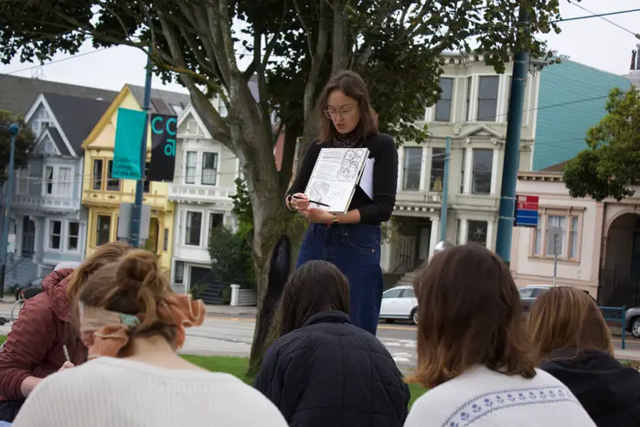 Natalie demonstrating a sketchbook drawing to workshop participants seated outdoors, with San Francisco houses behind her.