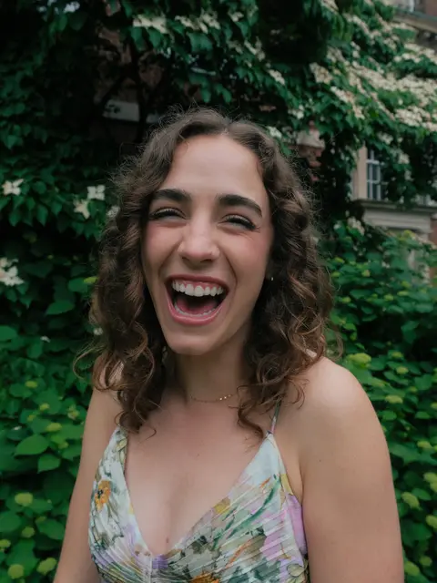 Close portrait of a wedding guest in a floral dress laughing with her eyes closed, framed by green leaves and white blossoms.