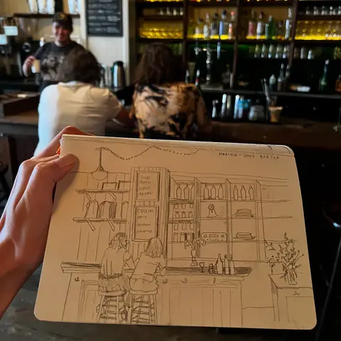 Natalie holding up her sketchbook drawing of the bar in front of the bar itself, mid-sketch