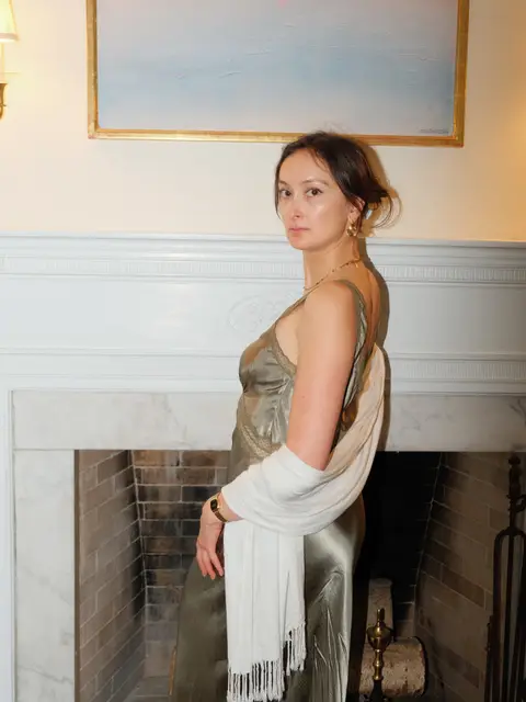 Quiet portrait of a wedding guest in a sage-green silk slip dress and white shawl, standing beside a marble fireplace under a framed seascape painting.