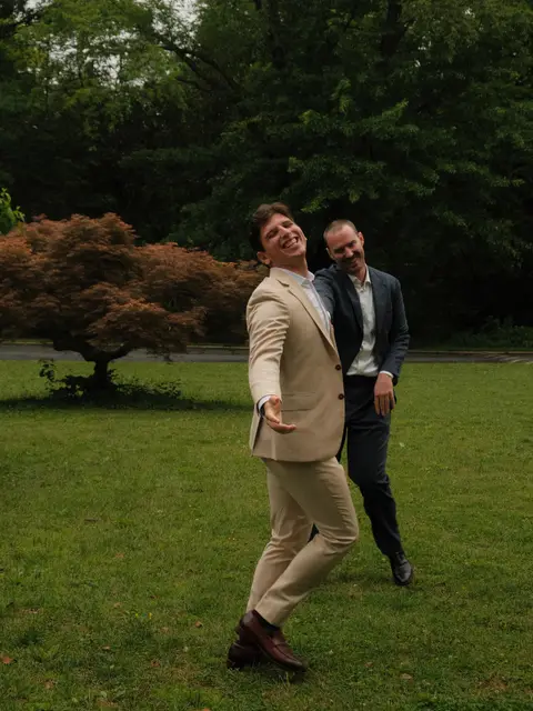 Two wedding guests in suits dancing and laughing on the lawn at the reception, a copper Japanese maple behind them.